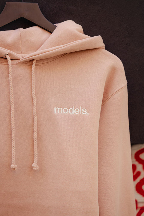 Detail picture of a Dusty pink hoodie with off white 'models.' embroidered logo on a hanger against a dark background