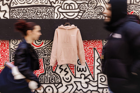 Pink hoodie on a hanger against a colorful, abstract wall covered with graffiti with people walking by.