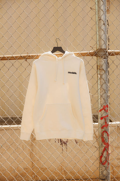 Off white hoodie with black embroidered 'models' logo on a hanger against a chain-link fence.