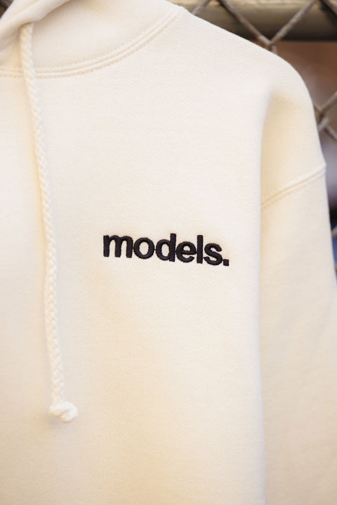 Off white hoodie with black embroidered 'models' logo  against a chain-link fence.