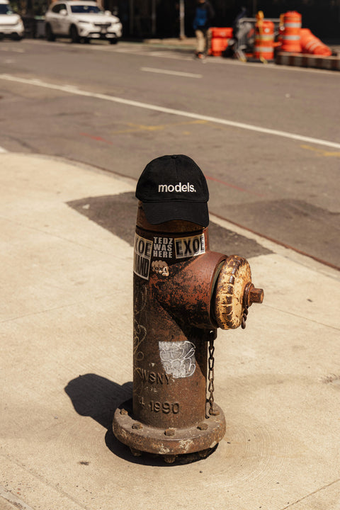Black cap with white 'models.' embroidery text on a rust-colored fire hydrant with stickers on a sidewalk