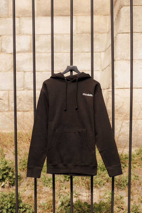 Black hoodie with a white"models." embroidered logo on the left side, on a hanger against a stone wall.