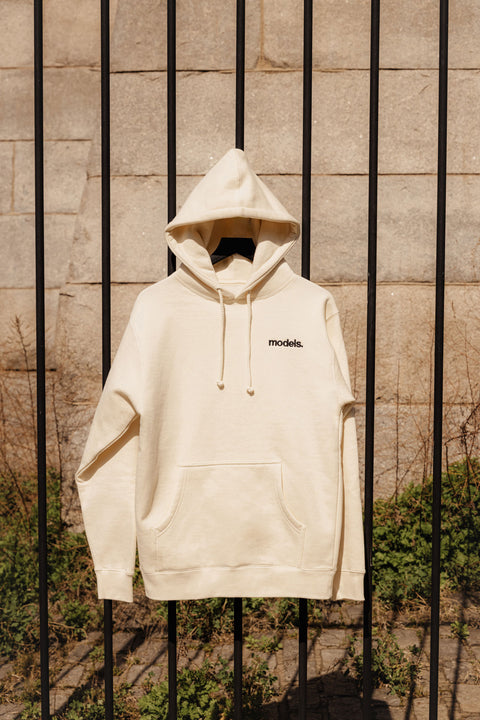Off white hoodie with black embroidered 'models' logo on a hanger against a metal fence.