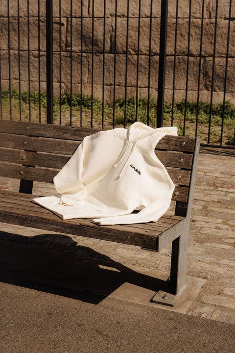 Off white hoodie with black embroidered 'models' logo on a wooden bench with a textured wall and grass in the background
