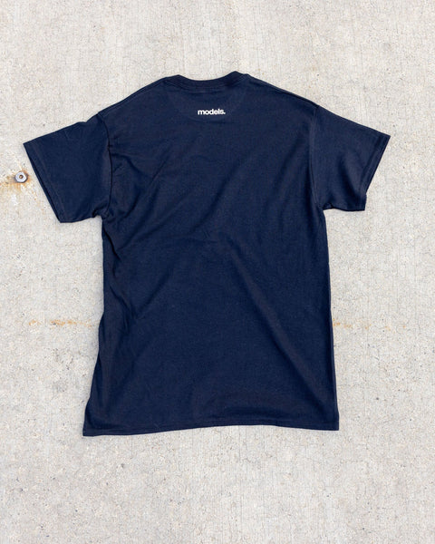 A black short-sleeve t-shirt laid flat on a textured light gray concrete surface. A small white 'Models.' brand logo is printed on the upper back of the shirt.