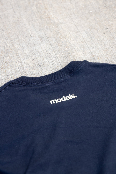 Close-up of the back collar of a black t-shirt with the word 'models.' printed in white lowercase letters, laid flat on a textured concrete surface.