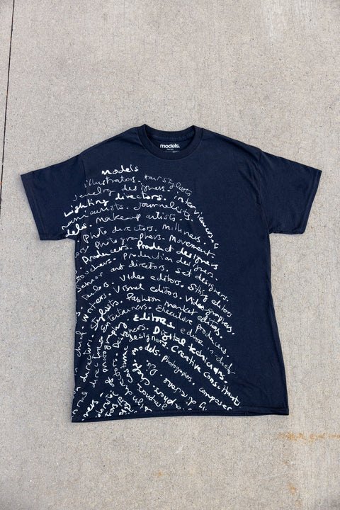 A black crewneck t-shirt with a white fingerprint pattern made of handwritten text, laid flat on a light gray concrete surface.