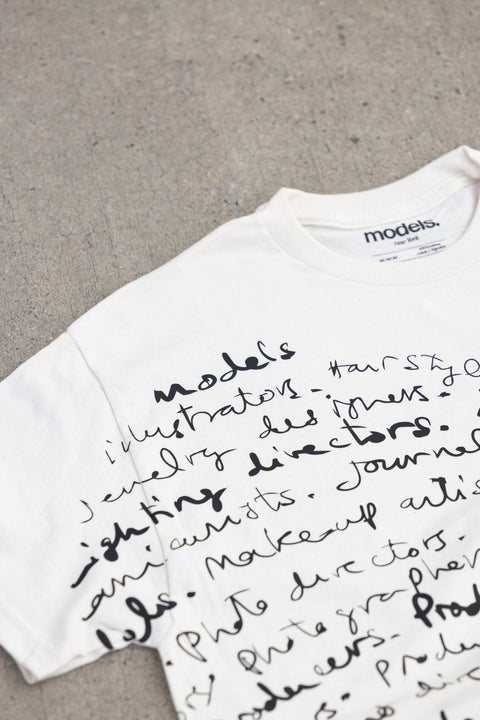 A white short-sleeve t-shirt with black handwritten-style text printed across the front, laid flat on a gray concrete surface. The inner neck label shows the brand 'models'.