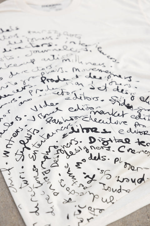 A white t-shirt featuring a dense, hand-written fingerprint style black graphic print of various words and phrases related to design, editing, and production, laid flat on a neutral gray surface.