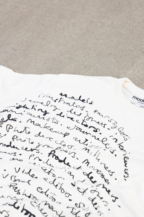 A close-up of a white crewneck t-shirt featuring a black handwritten-style graphic print of various professions, laid flat on a textured gray surface.