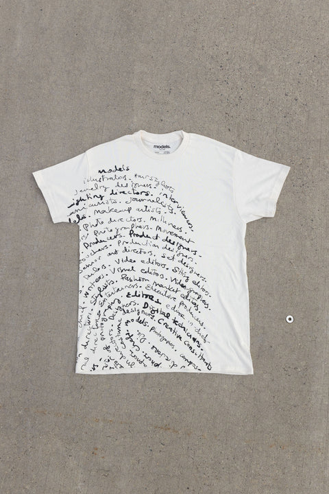 A white crewneck tee with a black fingerprint pattern made of handwritten text, laid flat on a light gray concrete surface.