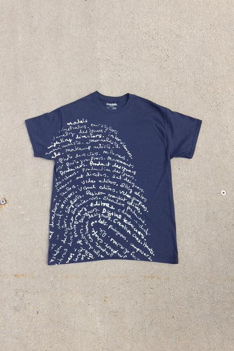 A navy blue short-sleeve t-shirt with a white script graphic resembling a fingerprint pattern, laid flat on a textured concrete surface.