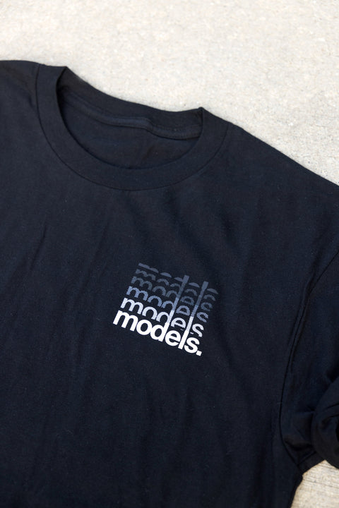 A close-up of a black t-shirt laid flat on a light gray textured surface. The shirt features a small graphic on the left chest with a gradient of the word 'models.'