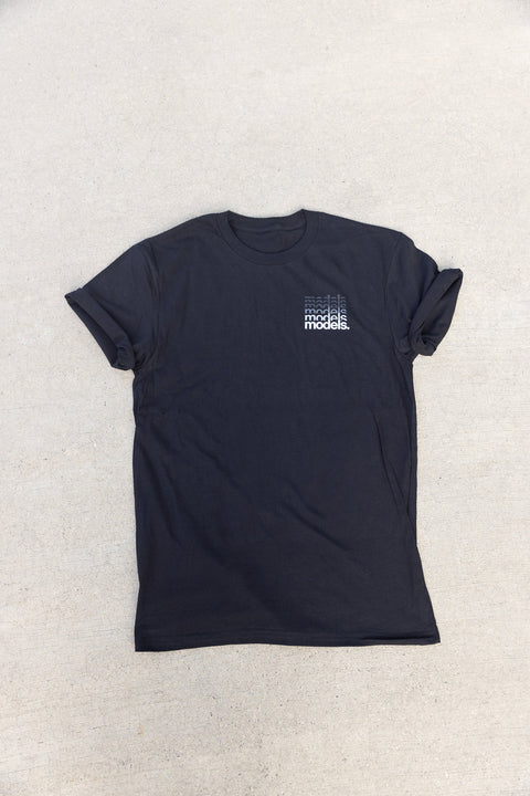 A black t-shirt styled with rolled sleeves, laid flat on a light gray textured surface. The shirt features a small graphic on the left chest with a gradient of the word 'models.'