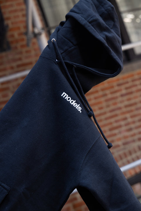 A close-up of a black pullover hoodie with a white embroidered 'models.' logo on the chest, featuring black drawstrings, set against a blurred brick wall background.