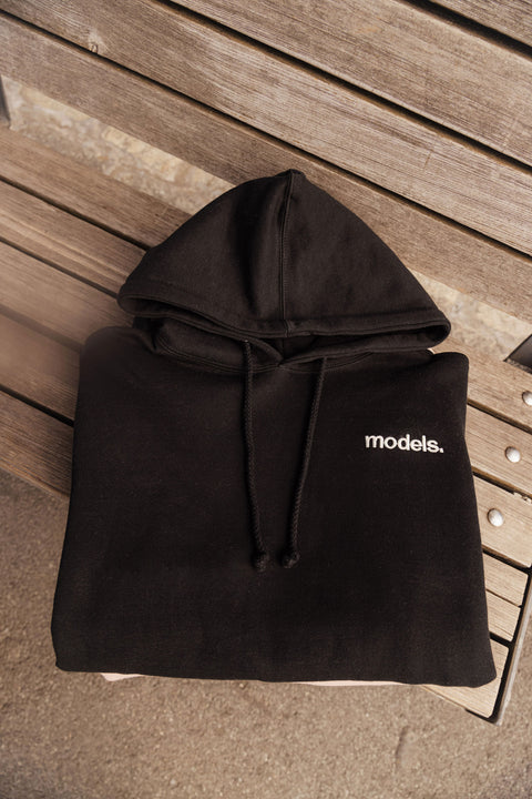 Folded black hoodie with white 'models.' embroidery  logo on a wooden bench surface