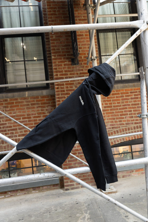 A black pullover hoodie with a white 'Models.' logo on the chest, draped over a metal scaffolding structure in front of a brick building.