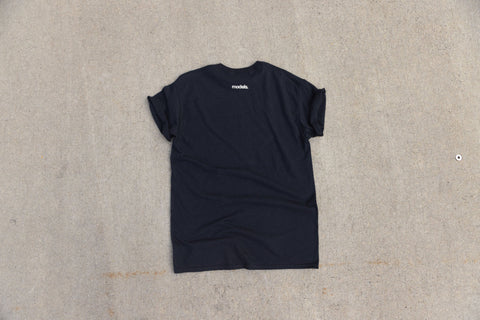 A black short-sleeve t-shirt with a small white 'models.' logo printed below the back neck, laid flat on a textured gray concrete surface.