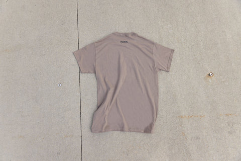 A short-sleeve crewneck t-shirt in a muted brown savana color, laid flat on a light gray concrete surface.