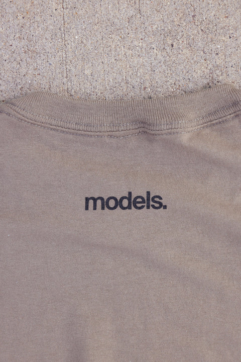 Close-up of a brown savana crewneck T-tshirt with the word 'models.' printed in black lowercase letters below the neckline, laid flat against a textured concrete background.
