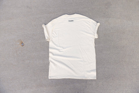A white short-sleeve t-shirt laid flat on a textured gray concrete surface, showing the back side with a small black 'models.' logo printed near the neckline.