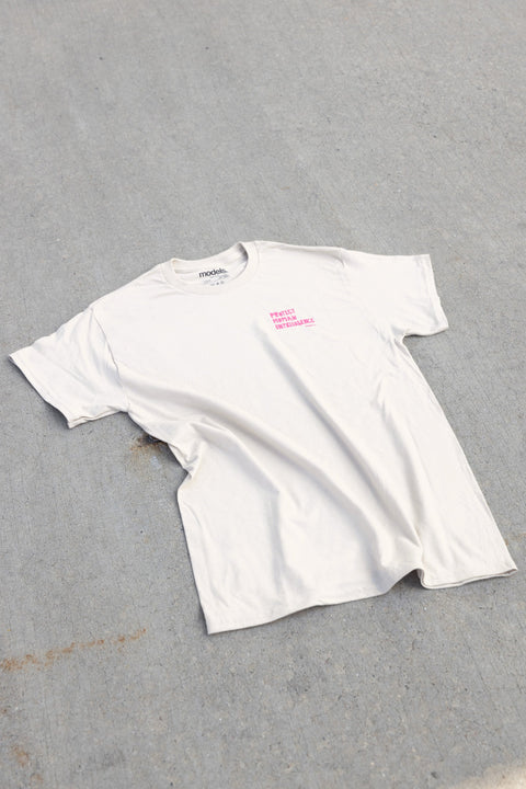 A white short-sleeve t-shirt with a small pink graphic on the left chest, laid flat on a gray concrete surface.