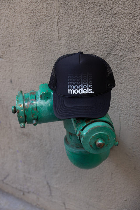 Front view of a black trucker hat with a white 'models.' gradiant logotype on the front panel in a white graphic gradient print resting on a distressed green outdoor hydrant against a beige stucco wall.