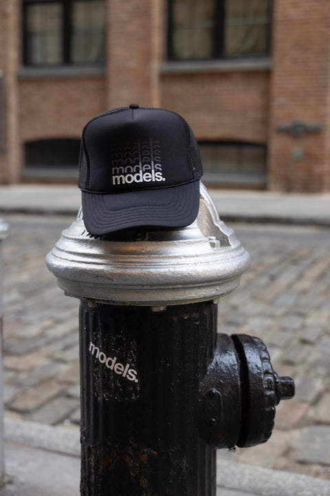 Front view of a black trucker hat with a white 'models.' gradiant logotype on the front panel resting on a fire hydrant against a cobble street.