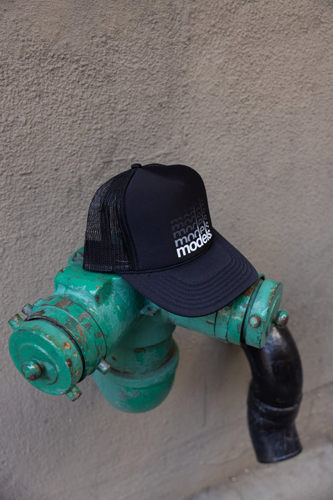 Side view of a black trucker hat with a white 'models.' gradiant logotype on the front panel in a white graphic gradient print resting on a distressed green outdoor hydrant against a beige stucco wall.