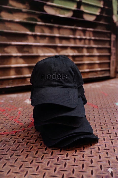 Black cap with black 'models.' embroidery text on a rusted landing dock surface.